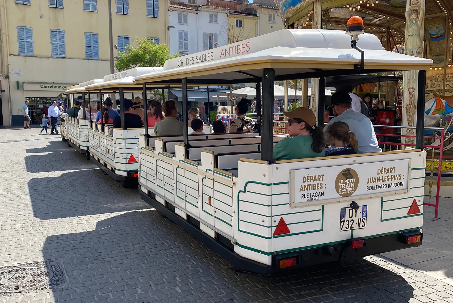 L’arrière du Petit Train d’Antibes et ses passagers sur une place, avec le panneau des départs d’Antibes rue Lacan et de Juan-les-Pins boulevard Baudoin